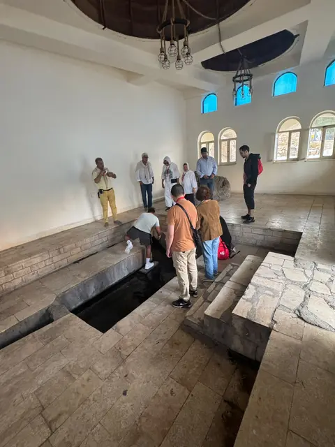 Groupe de visiteurs dans une salle historique avec puits anciens, fenêtres en arche et vitraux bleus.